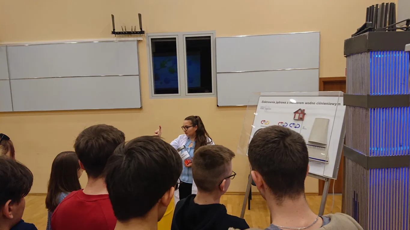 Edukator Fundacji objaśnia uczniom planszę o elektrowni jądrowej obok modelu kasety paliwowej