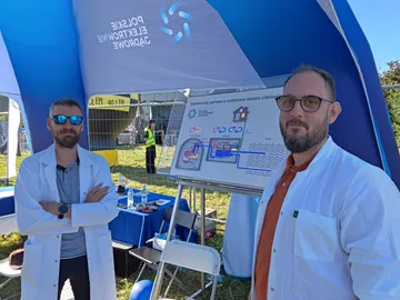 Dwie osoby w białych fartuchach przed planszą o elektrowni jądrowej z reaktorem wodno-ciśnieniowym