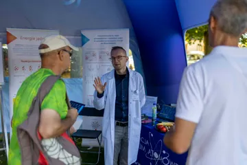 Osoba w białym fartuchu rozmawia ze zwiedzającymi przy planszach o energii jądrowej