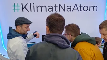 Osoba w białym fartuchu rozmawia ze zwiedzającymi przed ścianą z napisem #KlimatNaAtom