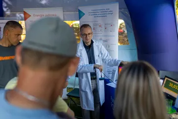 Osoba w białym fartuchu wskazuje eksponaty na stole, wokół stoją zwiedzający