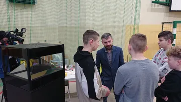 Osoba w marynarce rozmawia z grupą uczniów obok czarnej gabloty; w tle kamera telewizyjna