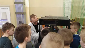 Osoba w białym fartuchu pokazuje dzieciom urządzenie w czarnej gablocie z szybą