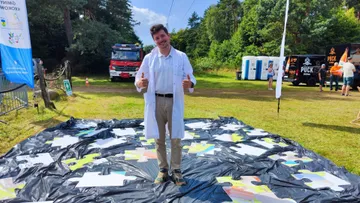 Osoba w białym fartuchu na rozłożonych na trawie dużych puzzlach; w tle wóz strażacki