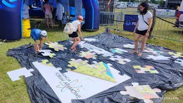 Dzieci i dorośli układają na trawie duże puzzle z logo Fundacji