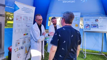 Dwie osoby w białych fartuchach rozmawiają ze zwiedzającym przy planszach o promieniowaniu