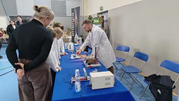 Osoba w białym fartuchu pokazuje doświadczenie przy stole zwiedzającym, w tym dzieciom
