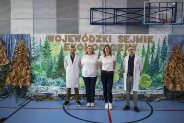 Cztery osoby, dwie w białych fartuchach, przed dekoracją z napisem „Wojewódzki Sejmik Ekologiczny”