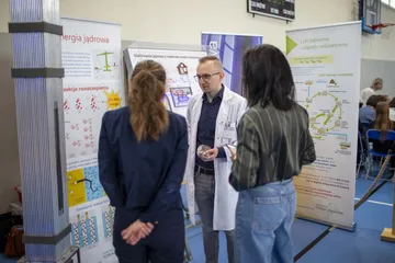 Osoba w białym fartuchu rozmawia z dwiema zwiedzającymi przy planszach o energii jądrowej i cyklu paliwowym