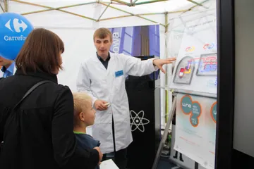Osoba w białym fartuchu wskazuje elementy planszy dziecku i dorosłym