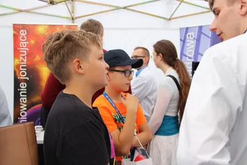 Dzieci słuchają osoby w białym fartuchu przy stoisku