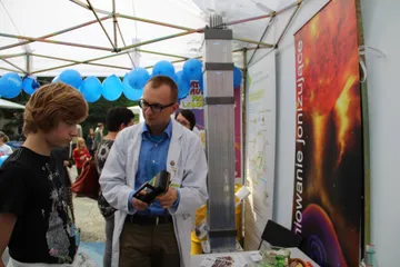 Osoba w białym fartuchu z miernikiem rozmawia z nastolatkiem przy stoisku