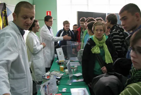 Osoby w białych fartuchach pokazują młodzieży doświadczenia przy stoisku
