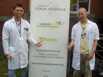 Dwie osoby w białych fartuchach przy roll-upie Fundacji FORUM ATOMOWE z logo projektów Spotkania z Energią Atomową, Nukleo i energiajadrowa.pl
