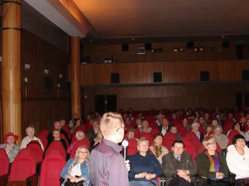 Prelegent z mikrofonem przed słuchaczami w sali z czerwonymi fotelami
