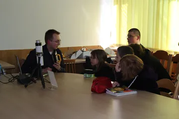 Osoba pokazuje młodzieży miernik przy stole; obok detektor na statywie