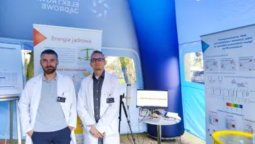 Dwie osoby w białych fartuchach przy stoisku z planszami o energii jądrowej w namiocie Polskich Elektrowni Jądrowych