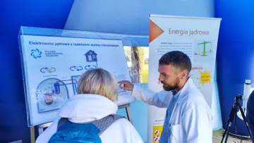 Osoba w białym fartuchu wskazuje zwiedzającej elementy planszy o elektrowni jądrowej