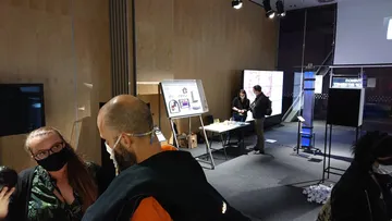 Dwie osoby w maseczkach rozmawiają na pierwszym planie; w tle stoisko z planszą i modelem kasety paliwowej