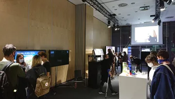 Zwiedzający w maseczkach przy stoisku Fundacji z ekranami w ciemnej hali Centrum Nauki Kopernik