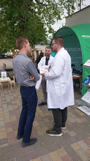 Dwie osoby w białych fartuchach rozmawiają ze zwiedzającym przed namiotem na placu