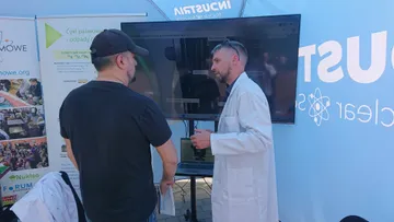 Osoba w białym fartuchu rozmawia ze zwiedzającym przed ekranem w namiocie Industria