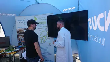 Osoba w białym fartuchu i zwiedzający przed ekranem przy planszy o cyklu paliwowym