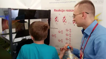 Osoba z Fundacji objaśnia chłopcu planszę o reakcji rozszczepienia