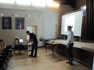 Osoba prowadząca wykład przy ekranie na scenie szkolnej auli; obok druga osoba przy laptopie