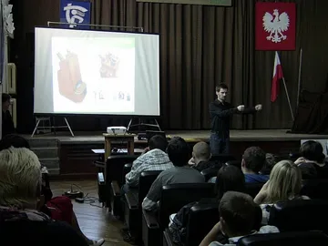 Wykład na scenie auli; na ekranie ilustracja przyrządów, na widowni uczniowie