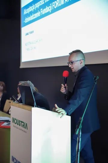 Osoba przemawia z mikrofonem przy mównicy Industria; za nią ekran z prezentacją Fundacji