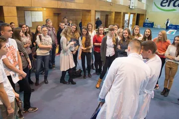 Dwie osoby w białych fartuchach przed kręgiem uczniów w sali gimnastycznej