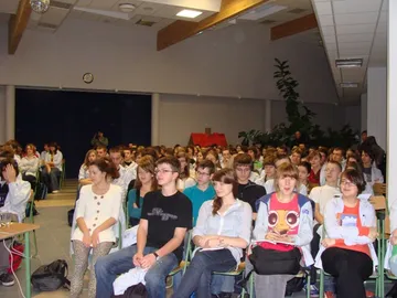 Uczniowie w białych fartuchach w rzędach krzeseł w auli