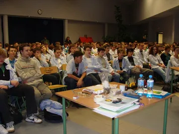 Aula pełna uczniów w białych fartuchach; na pierwszym planie stół z przyrządami do pokazów