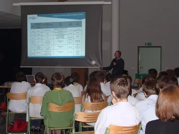 Wykład przy ekranie z tabelą o biologicznych skutkach promieniowania; na pierwszym planie uczniowie