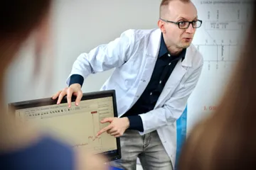 Osoba w białym fartuchu pokazuje uczniom tablicę