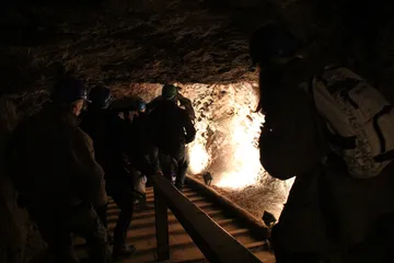 Zwiedzający w kaskach idą drewnianym pomostem w chodniku kopalni