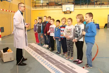 Osoba w białym fartuchu przy taśmie z widmem promieniowania przed rzędem uczniów
