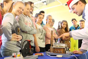 Uczniowie przy stole z maszyną parową i osobą w białym fartuchu
