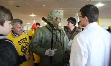 Protestujący w masce przeciwgazowej i osoba w żółtej koszulce z napisem „STOP” przy stoisku; obok osoba w białym fartuchu