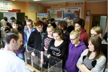 Uczniowie przy stoisku z eksponatami; obok osoba w białym fartuchu
