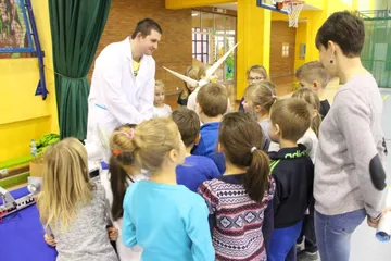 Osoba w białym fartuchu opowiada grupie dzieci przy makiecie