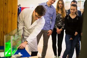 Osoba w białym fartuchu prowadzi doświadczenie z zielonym laserem i komorą z cieczą