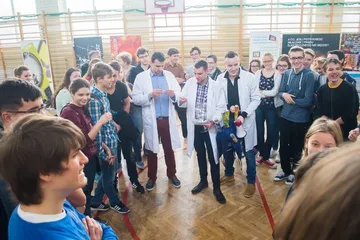 Kilka osób w białych fartuchach wśród uczniów w sali gimnastycznej