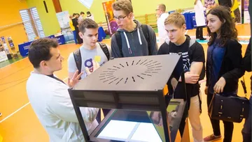 Osoba w białym fartuchu przy gablocie z hologramem; obok uczniowie