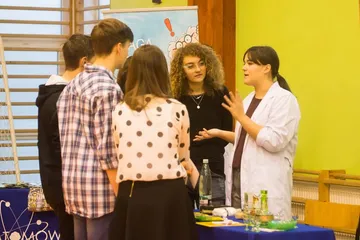 Osoba w białym fartuchu rozmawia z uczniami przy stole z doświadczeniami