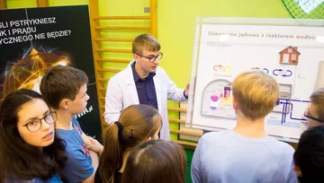 Osoba w białym fartuchu objaśnia uczniom podświetlany model elektrowni jądrowej