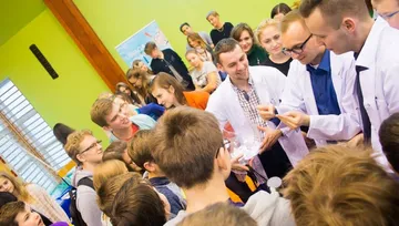 Kilka osób w białych fartuchach pokazuje doświadczenie grupie uczniów