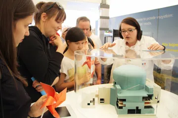 Osoba w białym fartuchu objaśnia dzieciom makietę reaktora