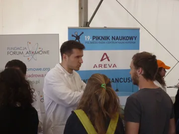 Osoba w białym fartuchu rozmawia ze zwiedzającymi przy roll-upie Pikniku Naukowego